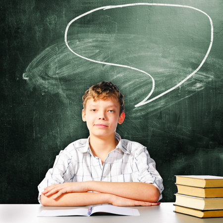 school boy is sitting at table with chalk board behind himの写真素材