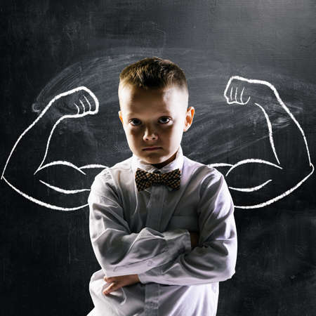 school boy is standing with strong hands on blackboard behind himの写真素材