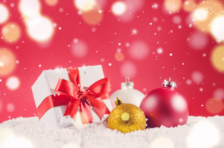 white decorative christmas gift box with ribbon and balls on snow against red festive backgroundの写真素材