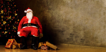 Tired Santa Claus sitting with gift boxes near decorated fir treeの写真素材