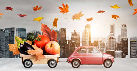 Food delivery. Autumn red toy car with fallen leaves delivering fruits and vegetables against business district buildingsの写真素材