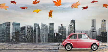 Autumn red toy car with fallen leaves against business district buildingsの写真素材