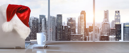 Laptop on table with Santa Claus hat in office with panoramic sunset view of modern downtown skyscrapers at business districtの写真素材