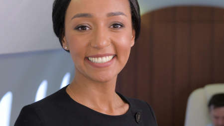 Stewardess portrait in private jet. Female biracial flight attendant smiling inside of business airplane cabin with passengers on background.の写真素材