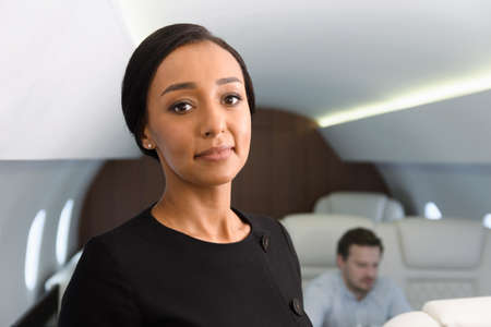 Stewardess portrait in private jet. Female biracial flight attendant smiling inside of business airplane cabin with passengers on background.の写真素材