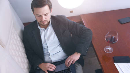 Business man working comfortably on laptop on board of private jet. Caucasian businessman travel inside of business airplane cabinwith glass of wine, view from above.の写真素材