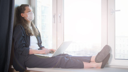 Bored woman looking at window and working on laptop from home due to coronavirus self isolation. Sitting on window sill. Authentic home workplace. Coronavirus outbreak 2020.の写真素材