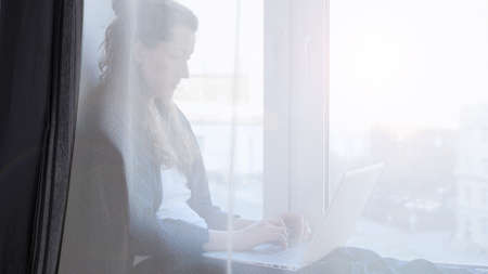Woman working on laptop from home due to coronavirus self isolation. Sitting on window sill. Authentic home workplace. Coronavirus outbreak 2020.の写真素材