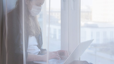 Woman wearing mask and working on laptop from home due to coronavirus self isolation. Sitting on...