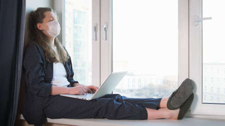 Bored woman looking at window and working on laptop from home due to coronavirus self isolation. Sitting on window sill. Authentic home workplace. Coronavirus outbreak 2020.の写真素材