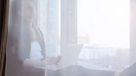Bored woman working on laptop from home due to coronavirus self isolation. Sitting on window sill. Authentic home workplace. Coronavirus outbreak 2020.の写真素材