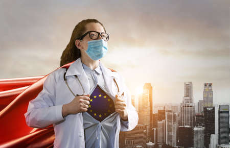 Medical superhero showing flag of EU. Heroic doctor wearing mask and red cape fighting with epidemic in city. Young woman. Brave medical staff of European Union concept.の写真素材