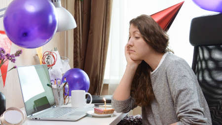 Sad single woman celebrating her birthday through video call virtual party. Sitting alone with laptop and cake with candle. Authentic workplace. Coronavirus outbreak 2020.の写真素材