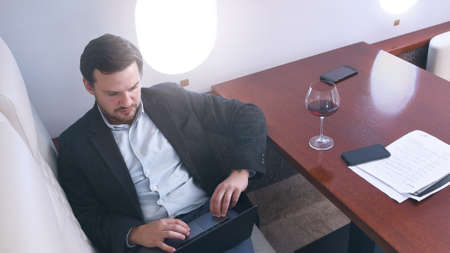 Business man working comfortably on laptop on board of private jet. Caucasian businessman travel inside of business airplane cabinwith glass of wine, view from above.の写真素材