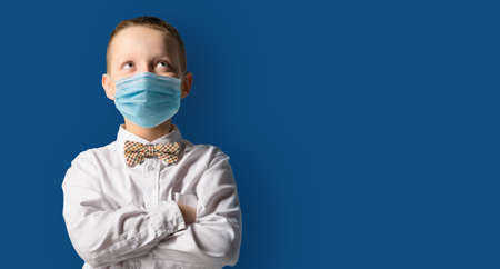 School boy wearing face mask standing against blackboard. Safe back to school during pandemic concept. New normal education. Hands folded and looking upwardsの写真素材