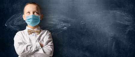 School boy wearing face mask standing against blackboard. Safe back to school during pandemic concept. New normal education. Hands folded and looking upwardsの写真素材