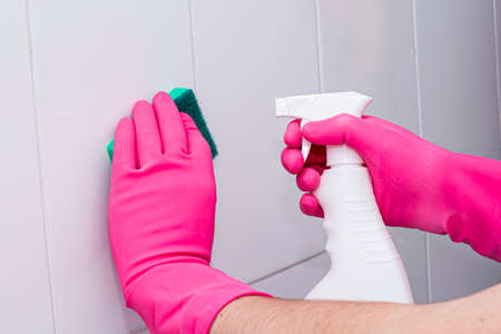 man in rubber household gloves with a sponge and spray washes a white tiled wall in the bathroomの写真素材