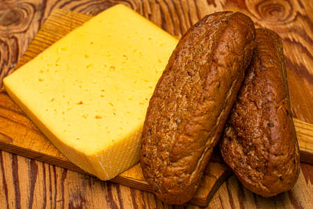 a piece of cheese and two rye bread rolls on a cutting board on a wooden table Rustic collection. Organic food. Organic product. High quality photoの写真素材