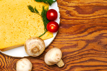 a piece of cheese greens cherry tomatoes and mushrooms on a plate on a wooden table copy space. High quality photoの写真素材