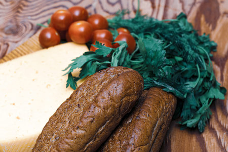 a piece of cheese greens cherry tomatoes and rye rolls on a wooden cutting board on the table. High...