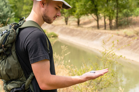 a tourist guy with a hat and a backpack is guided by a compass standing on the river bank. High...