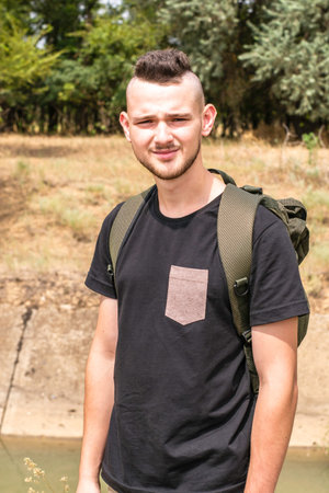 a tourist guy with a fashionable hairstyle and a backpack is standing on the river bank. High...
