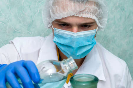 a medical laboratory assistant in a white coat gloves a hygienic mask and a cap pours liquid from one vessel to another of a larger volume. High quality photoの写真素材