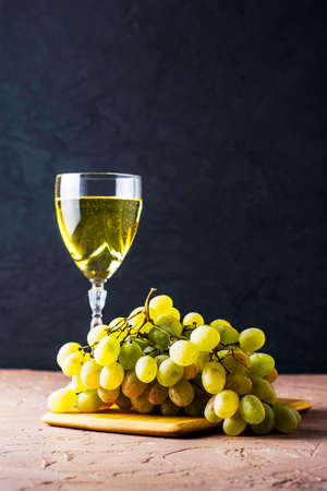 a brush of white grapes on a cutting board in the background a glass of white wine on a black background. High quality photoの写真素材