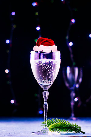 a glass of champagne with an ornament in the form of a Santa Claus hat and a fir twig near the leg on a black background with a backlight and another glass in the background. High quality photoの写真素材