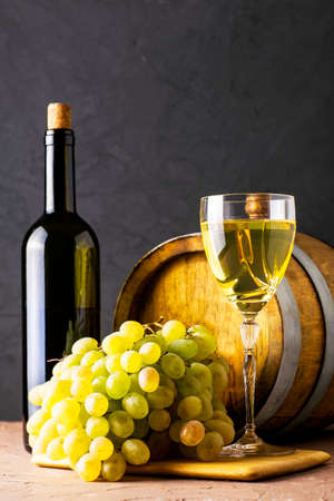 a bottle of wine a glass an oak barrel and a bunch of white grapes on a dark background. High quality photoの写真素材