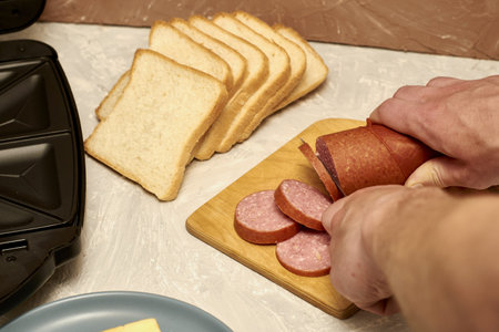a man prepares sandwiches in a sandwich maker cuts sausage with a knife on a wooden cutting board. high quality photoの写真素材