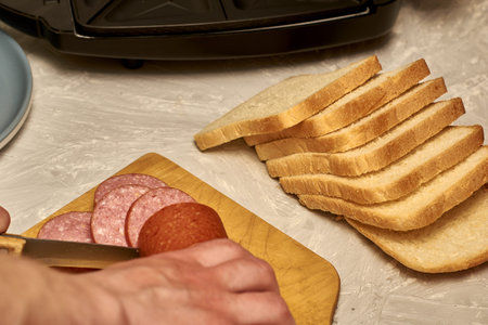 a man prepares sandwiches in a sandwich maker cuts sausage with a knife on a wooden cutting board