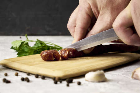 a man cuts spicy smoked sausages on a cutting board next to red pepper garlic spices and herbs on the table. high quality photoの写真素材