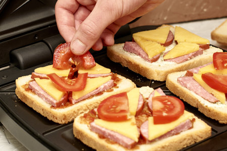 a man cooking sandwiches in a sandwich maker puts slices of tomatoes on bread. high quality photoの写真素材