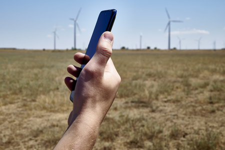 a man holds a smartphone in his hand against the background of a modern wind farm. mobile phone...