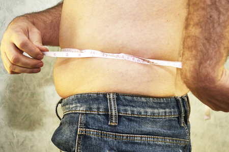 a man in jeans with a small belly measures the waist with a ribbon close-up - the concept of body positiveの写真素材