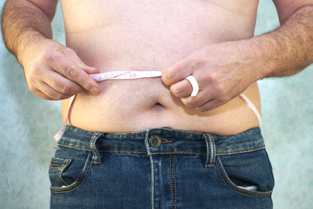 a man in jeans with a small belly measures the waist with a ribbon close-up - the concept of body positiveの写真素材