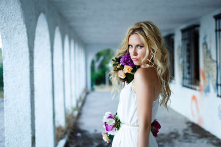 Young gorgeous blond Caucasian girl with blue eyes and long curly hair standing sideways with flowers tied to her body and white columns and a gate in the background.の写真素材