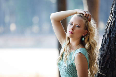 Young blond woman with long curly hair and blue eyes in the forest with a blurry background of a lake.  Beauty girl wearing a short green summer dressの写真素材