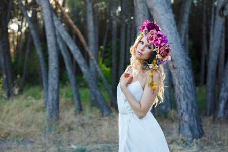 blond young woman with wavy long hair posing in romantic style in the forest. beautiful bride wearing elegant white dress and flower crown hairbandの写真素材
