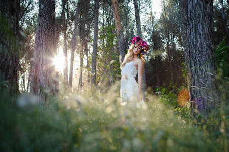Blond Caucasian woman with wavy hair, posing in romantic style in the forest. Young beautiful bride wearing elegant white dress, flowers and backlightの写真素材