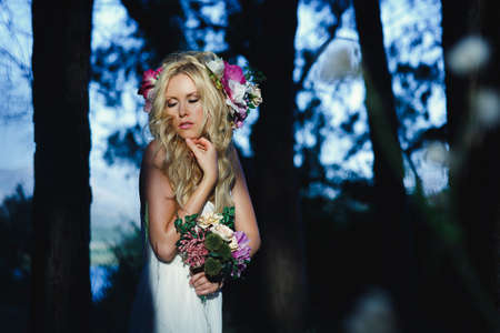 Beauty woman with long hair and flower hairband holding a bouquet. Bride posing in romantic style iluminated by sunlight  with her eyes closedの写真素材