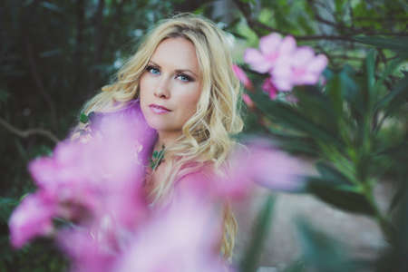 Bucolic portrait of young beautiful blond Caucasian female with blue eyes  surrounded by pink flowers and green leaves in the countrysideの写真素材