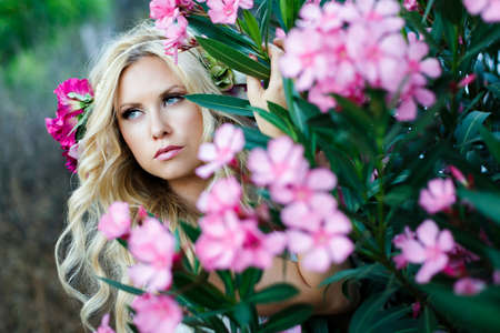 Bucolic portrait of young beautiful blond Caucasian female with blue eyes and wavy long hair surrounded by pink flowers and green leaves in forestの写真素材