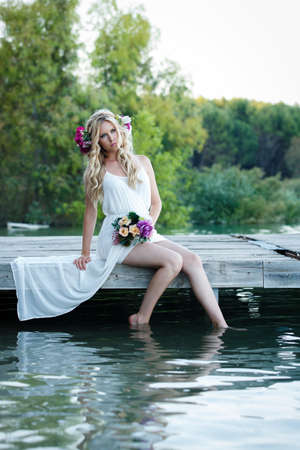 Attractive blond woman sitting in pier next to the lake with nature background. Romantic bride wearing white dress and holding flower bouquetの写真素材
