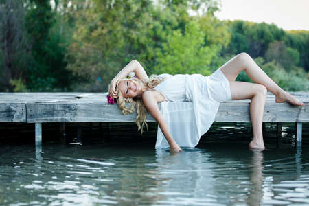Beautiful blond woman with long wavy hair wearing white dress with blurry nature background. Romantic bride lying on the wooden pier next to the lakeの写真素材