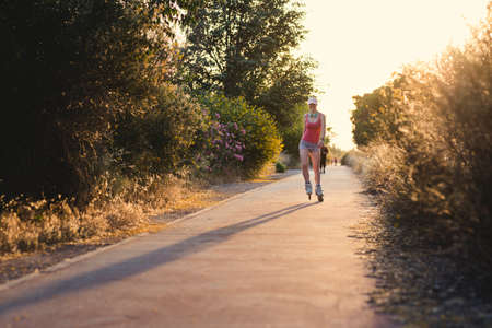 Young woman having fun while roller skating outdoors during sunset. Attractive female wearing a pale pink cap, headphones and sports clothes doing exercise.の写真素材
