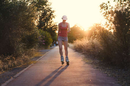 Young woman having fun while roller skating outdoors during sunset. Attractive female wearing a pale pink cap and sports clothes doing exercise with her headphones on listening to the music.の写真素材