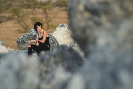 Beautiful young woman wearing black clothes and short haircut sitting on the rocks outdoors. Caucasian brunette girl smiling happy in the countryside.の写真素材