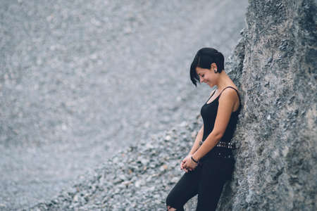 Caucasian young woman wearing black clothes smiling. Happy beautiful brunette girl leaning against gray rock wall outdoors.の写真素材